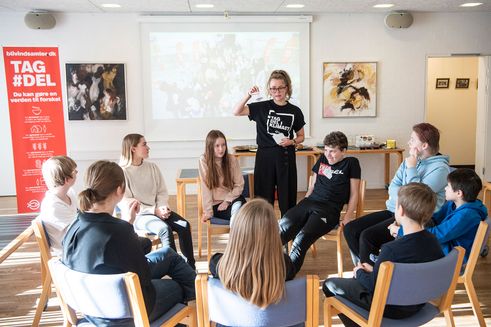 Ung kvinde fra TagDel-Karavanen underviser andre unge, der sidder i rundkreds rundt om hende på stole